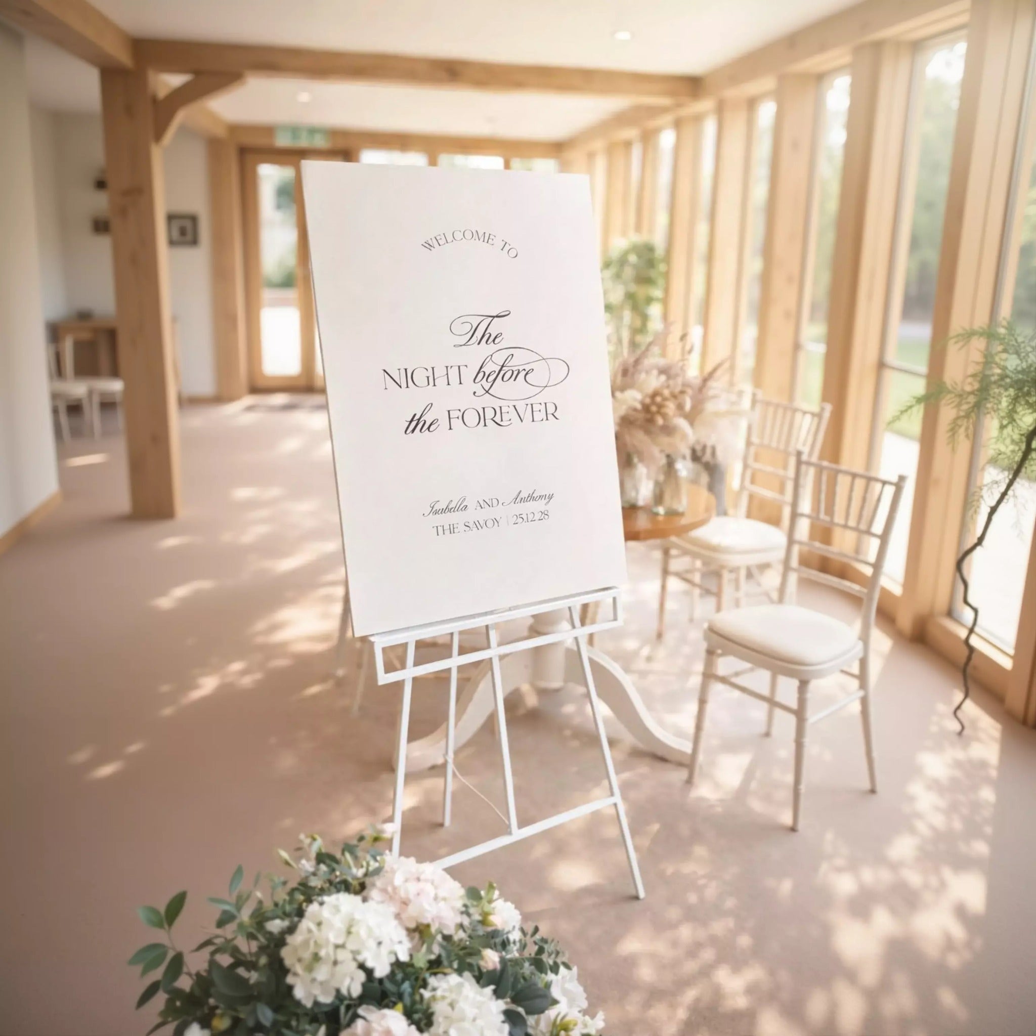 a white sign with elegant text that reads "Welcome to The Night Before the Forever" and "Isabella and Anthony The Swan 21/2/25". The sign is placed in a room with wooden beams and chairs, and there is a vase of flowers nearby.