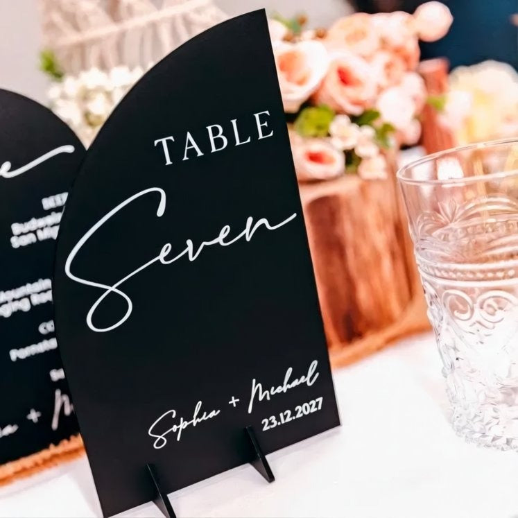 A black table sign displaying the text "TABLE Seven" and the names "Sophia & Michael" is placed on a table with a glass of water in the background.