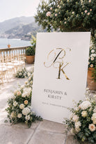 a white sign with a large "B" and "K" in gold lettering, surrounded by white flowers and greenery. The sign displays the names "Benjamin & Kirsty" and the date "18.03.28" at the bottom.