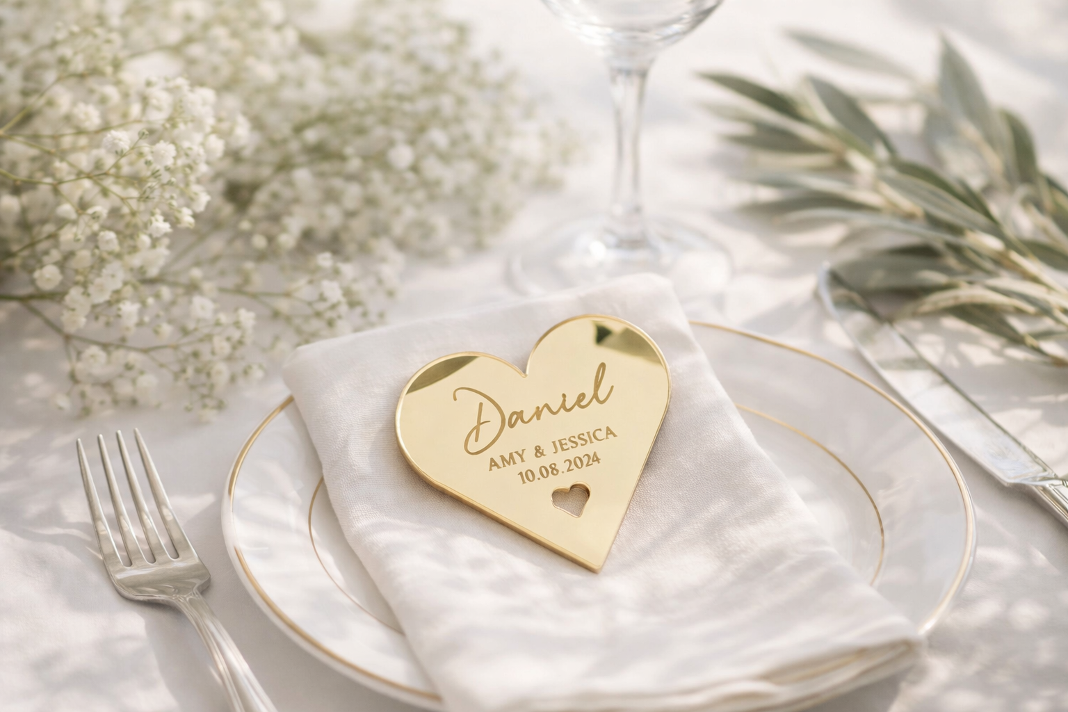 a heart-shaped place card with the names 'Daniel' and 'JESSICA' engraved on it, placed on a white tablecloth. The card is accompanied by a fork, knife, and a glass of wine, creating a romantic and elegant setting.