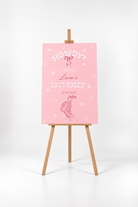 A pink poster with the text "HOWDY! Louisa's LAST RODEO! 30.03.2027" displayed on it, standing on a wooden easel.