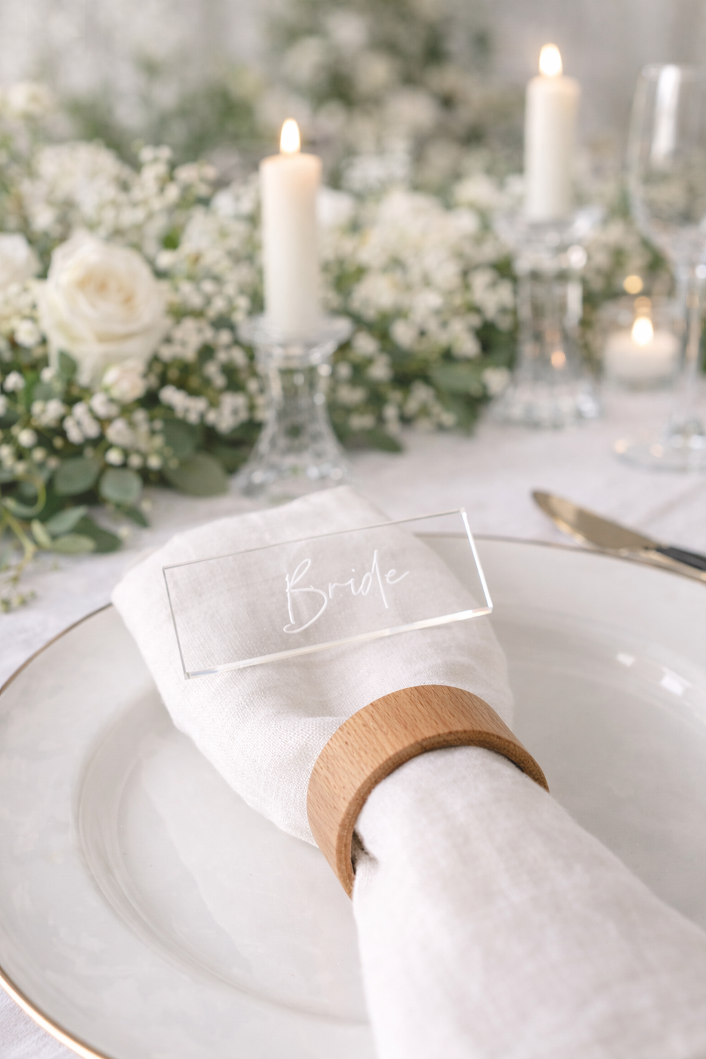 A table setting with a clear napkin holder displaying the word "Bride" and a wooden ring around a white napkin. The table is decorated with white flowers and candles, creating a romantic and elegant atmosphere.