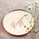 a round, reflective surface with the name "Reggie" written on it in cursive script. The surface is placed on a woven material, and there are small white flowers visible in the background.
