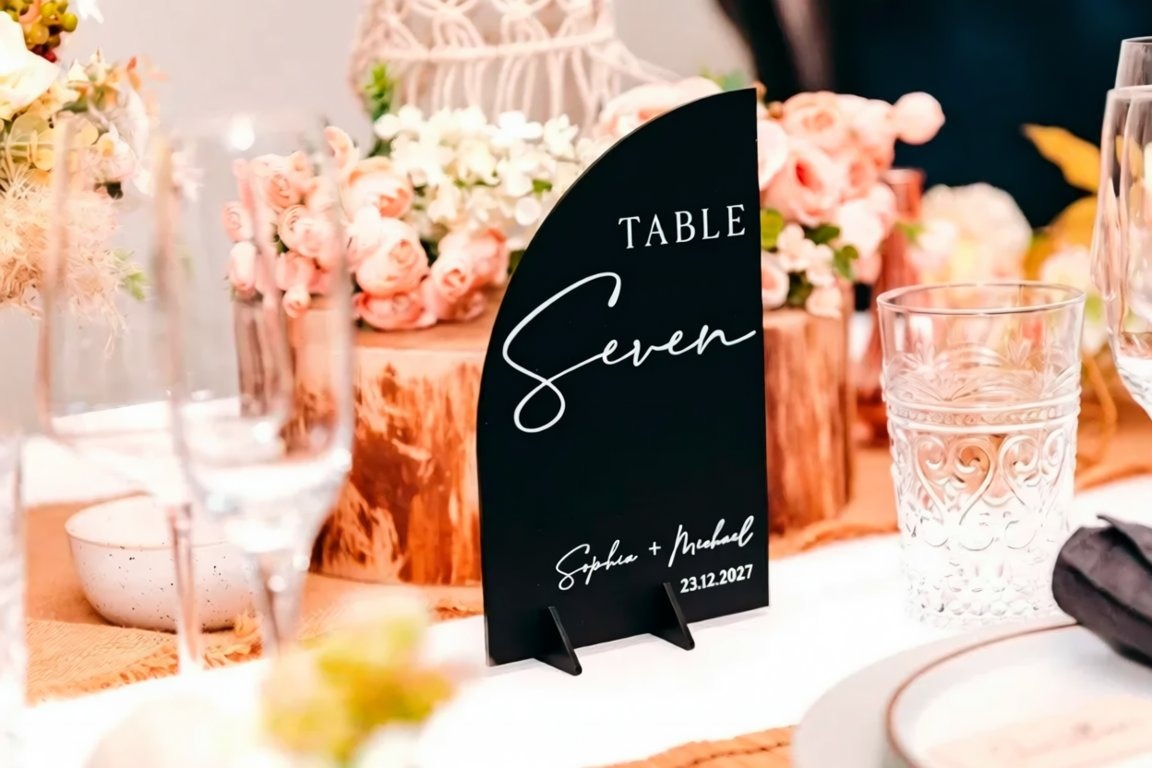 A table setting with a black table number sign displaying "TABLE Seven" and the names "Sophia & Michael" written on it, along with the date "23.12.2007". The table is decorated with pink and white flowers, and there are wine glasses and other tableware items visible.