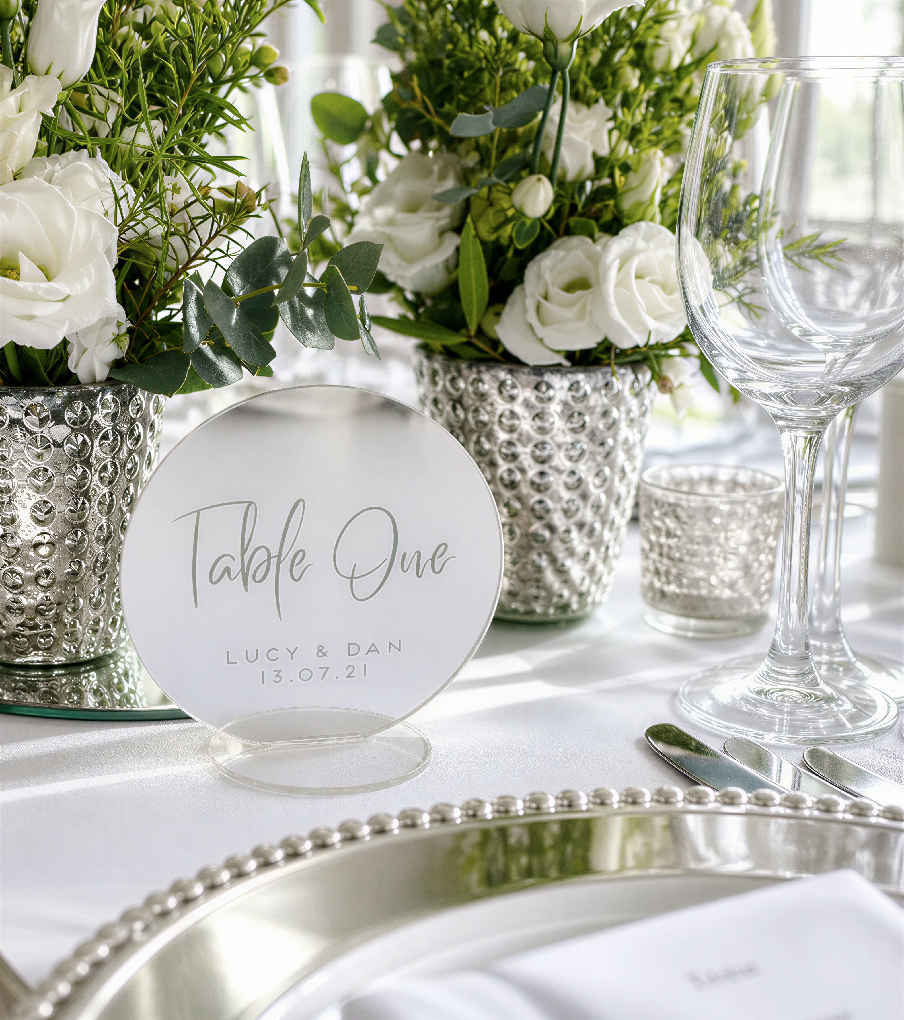a table setting with a table number sign, silverware, and a vase of white flowers.