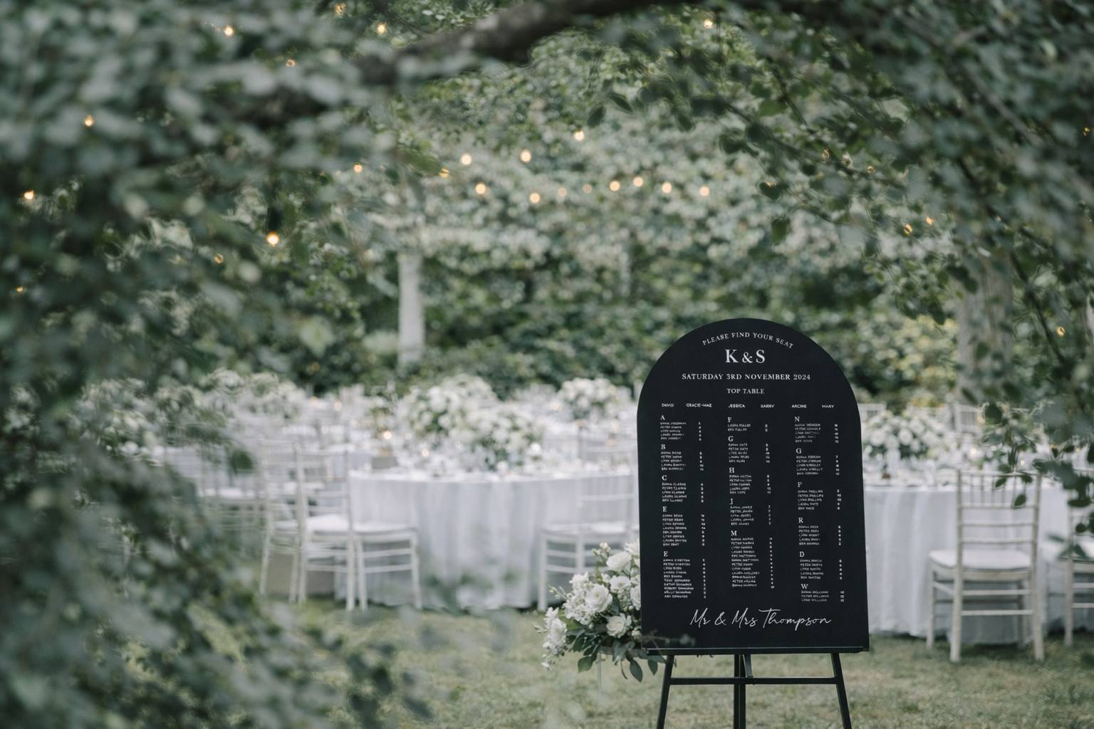 a black sign with white text, standing in a garden setting with tables and chairs arranged in the background.