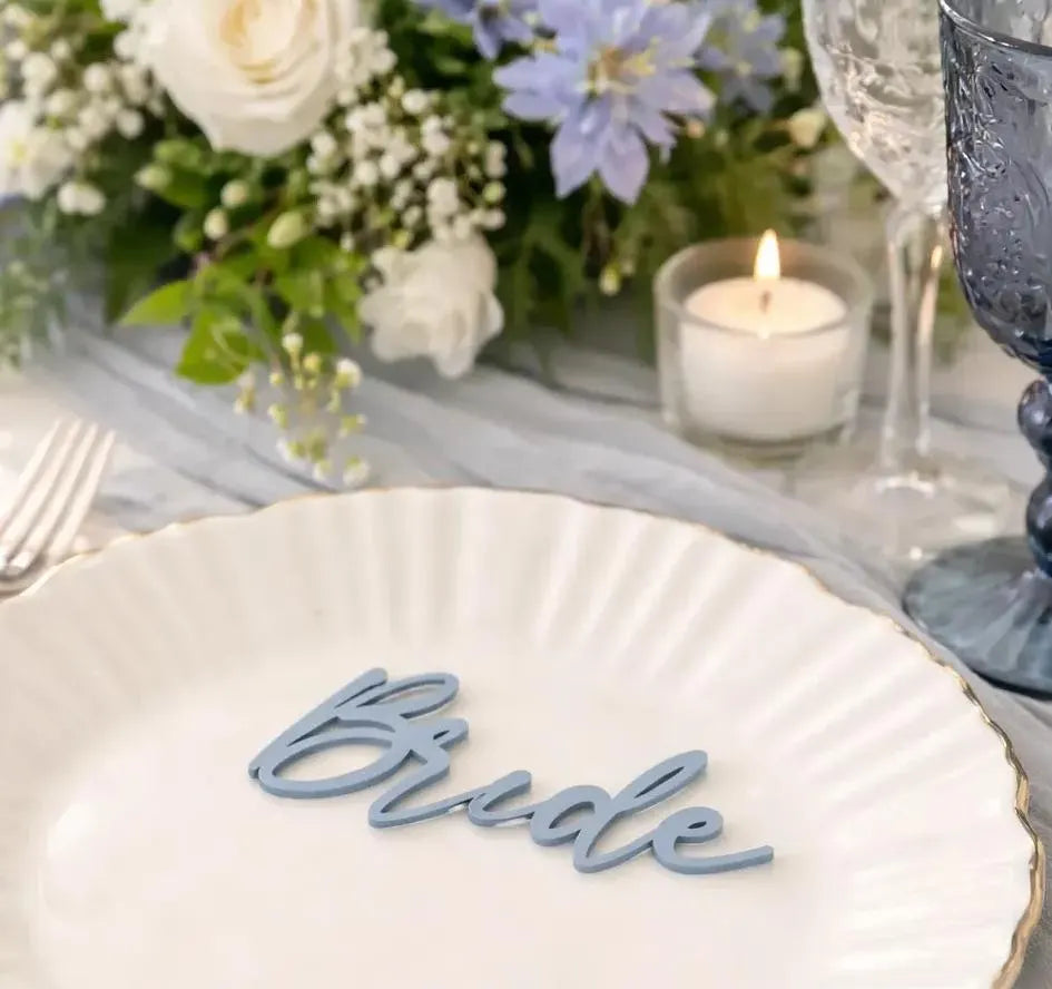 a table setting with a white plate, a candle, and a floral arrangement. The word "Bride" is written on the plate, and there are wine glasses and a vase of flowers in the background.