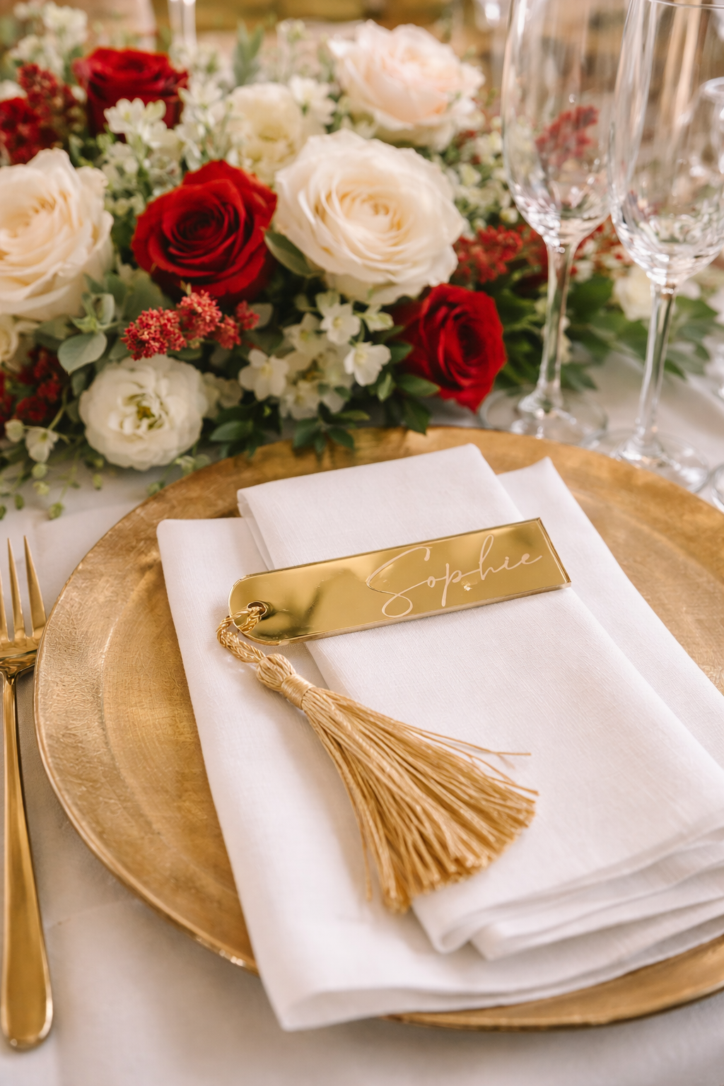 a beautifully set table with a gold plate, a white napkin, and a gold tassel. The table is adorned with a floral centerpiece and two wine glasses.