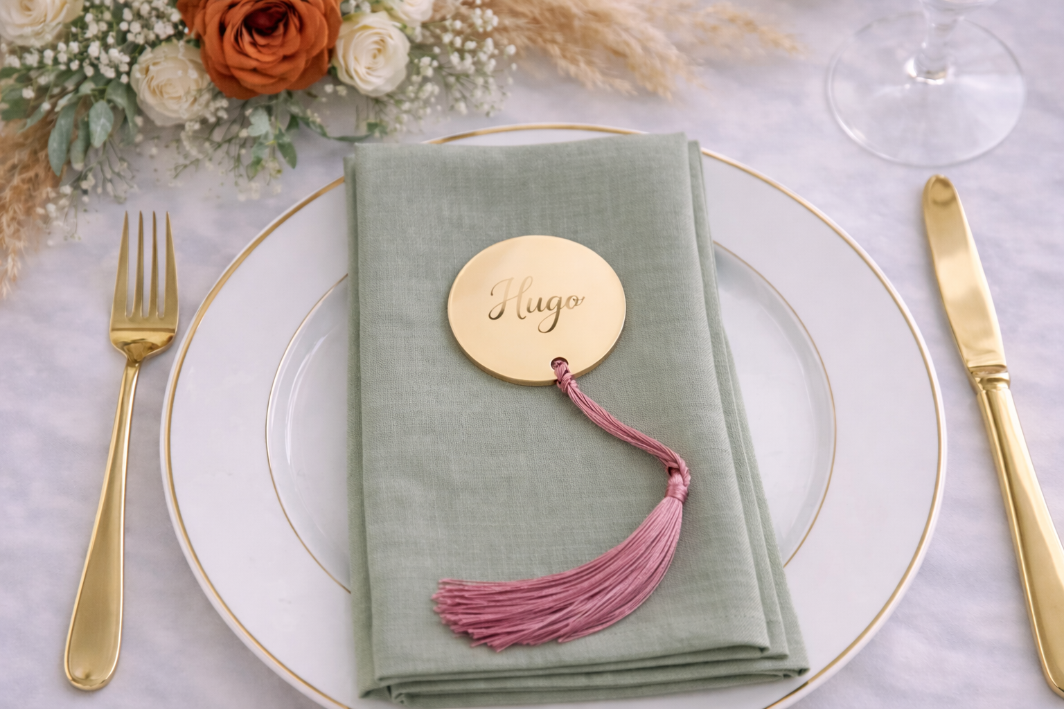 a table setting with a white plate, a green napkin, a gold tassel, and a small gold-colored name tag that says "Hugs". There are also a fork and a knife placed on the table.