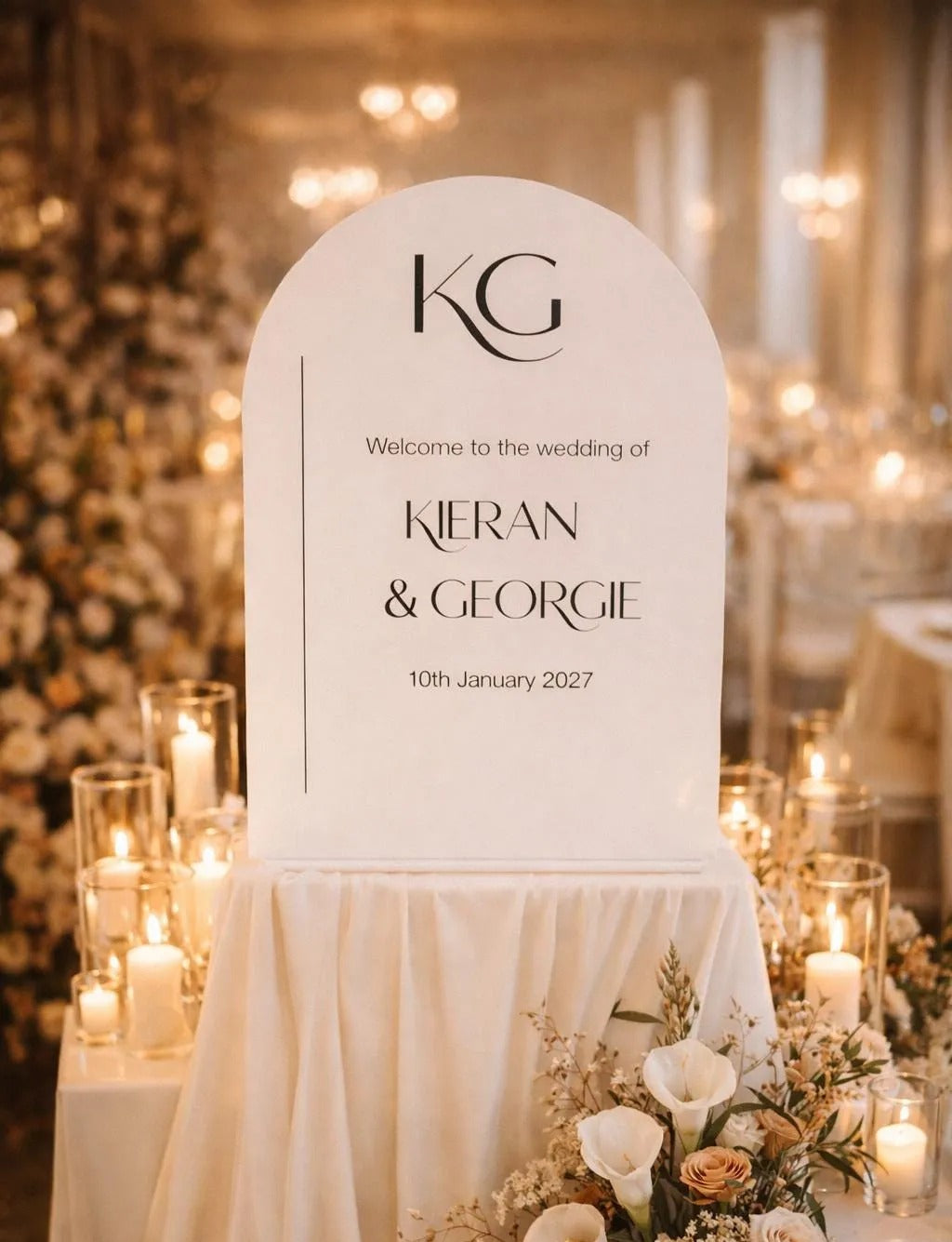 a wedding reception scene with a large white sign that reads "Welcome to the wedding of KIERAN & GEORGIE 10th January 2027". The sign is surrounded by a beautiful floral arrangement and lit candles, creating a warm and inviting atmosphere.