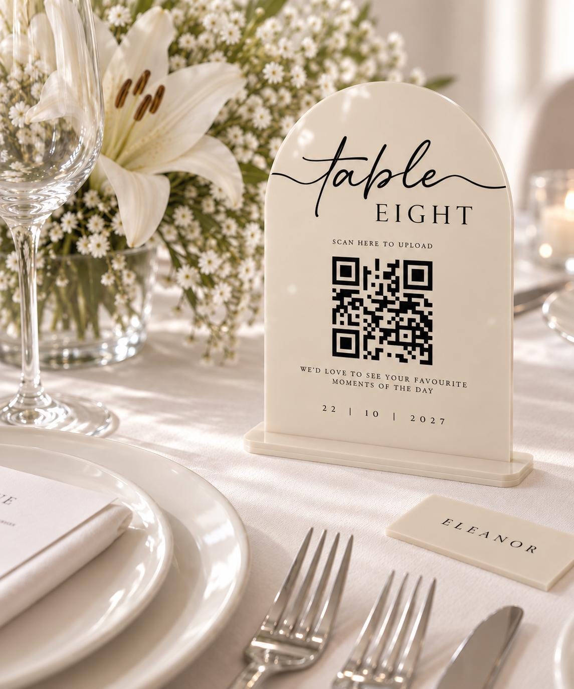 a table setting with a white tablecloth, plates, silverware, and a centerpiece of white flowers. There is a sign on the table that reads "table eight" and a QR code, suggesting a digital or electronic payment method.