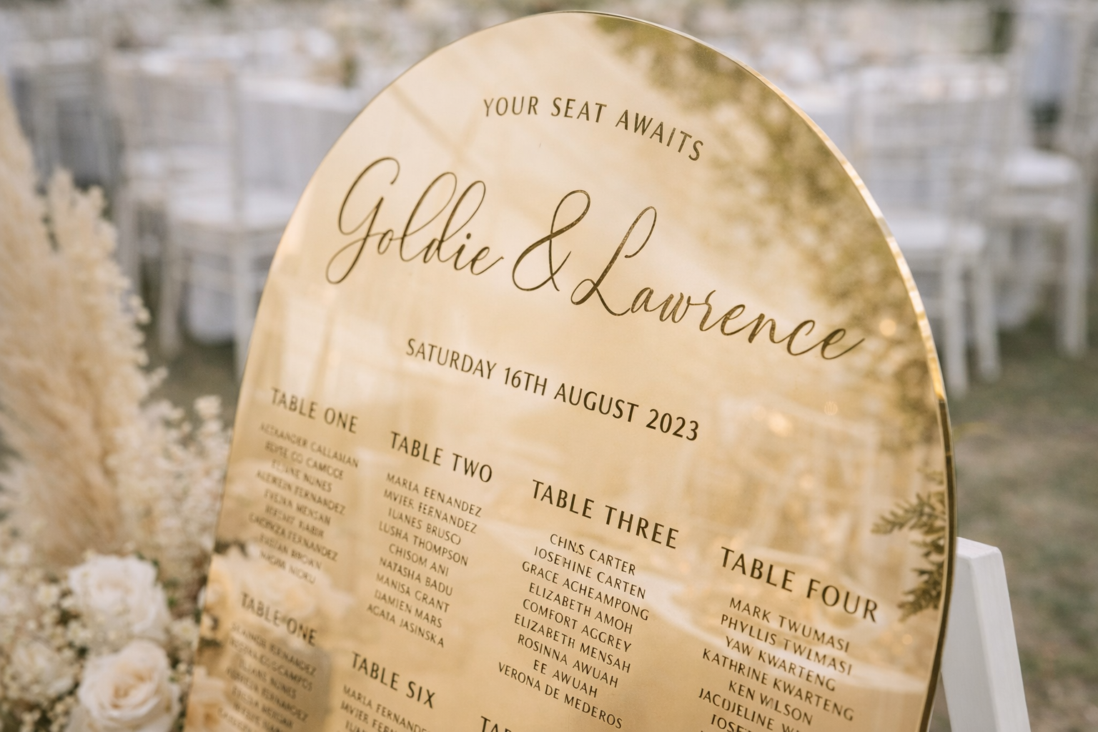 A close-up of a gold-colored sign displaying seating arrangements for an event, with the names of the guests and the date of the event.
