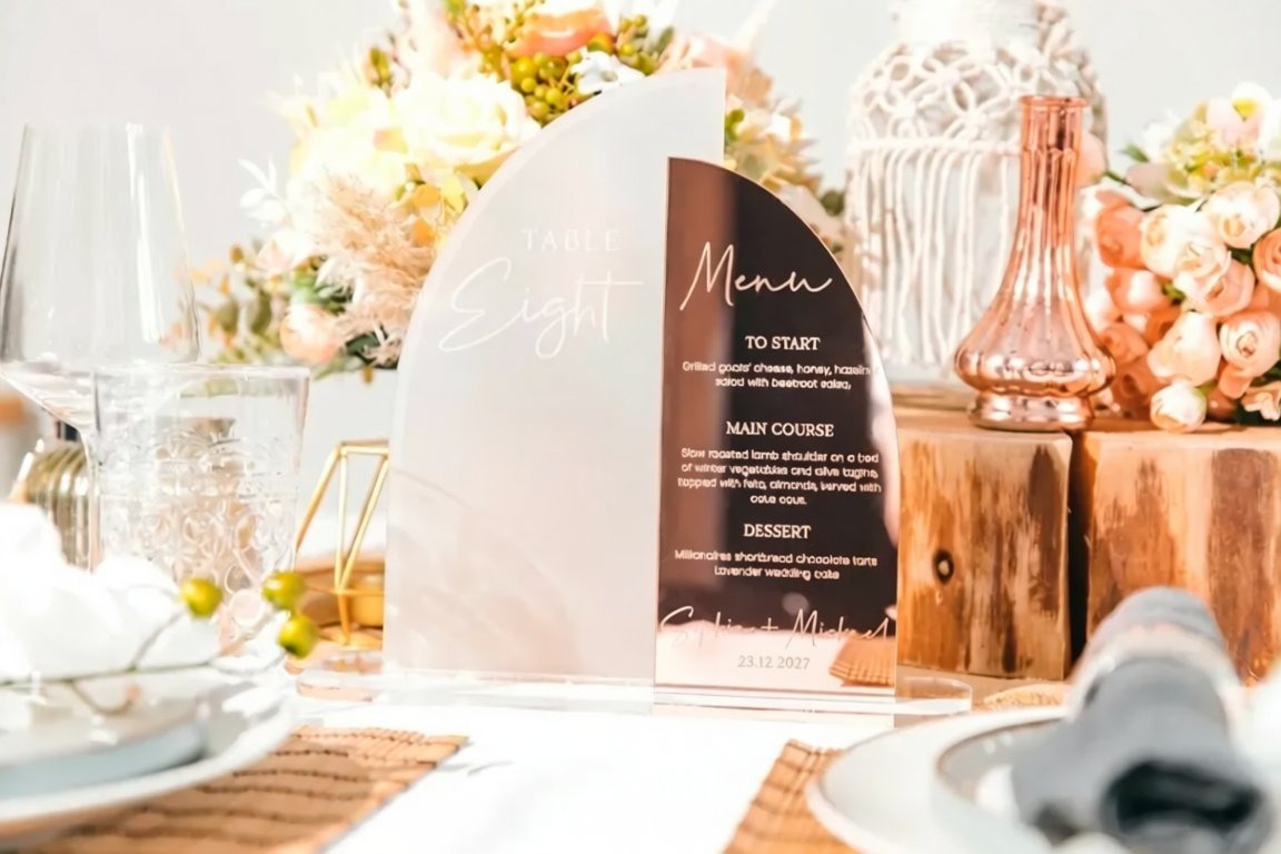 a table setting with a clear glass menu stand displaying a menu for the "Table Eight" course. The table is set with various tableware items, including wine glasses, cups, and a vase with flowers. The overall scene appears to be a formal dining setting, possibly for a wedding or other special event.