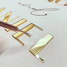 a close-up view of a person's hand using a tool to apply a clear adhesive to a piece of gold-colored material, which appears to be a decorative element.
