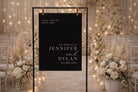 a black sign with white text, hanging in a room decorated with white flowers and lights, indicating a wedding ceremony.