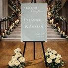 a wedding invitation sign with the names "Ella & Adrian" and the date "07.05.2026" displayed on it, standing on a wooden floor in front of a staircase decorated with flowers.