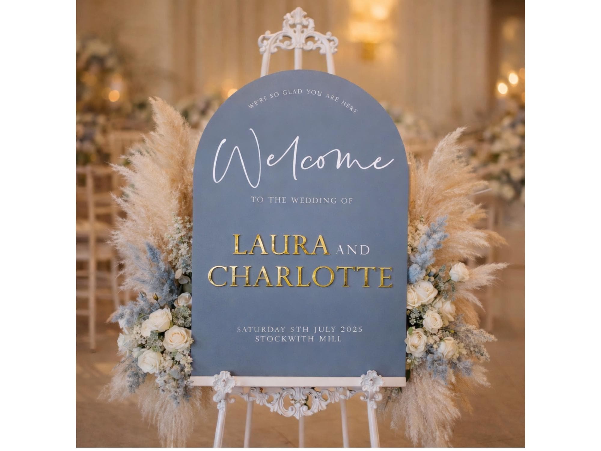 A blue wedding welcome sign with gold lettering, surrounded by pampas grass and white flowers, indicating the wedding of Laura and Charlotte.
