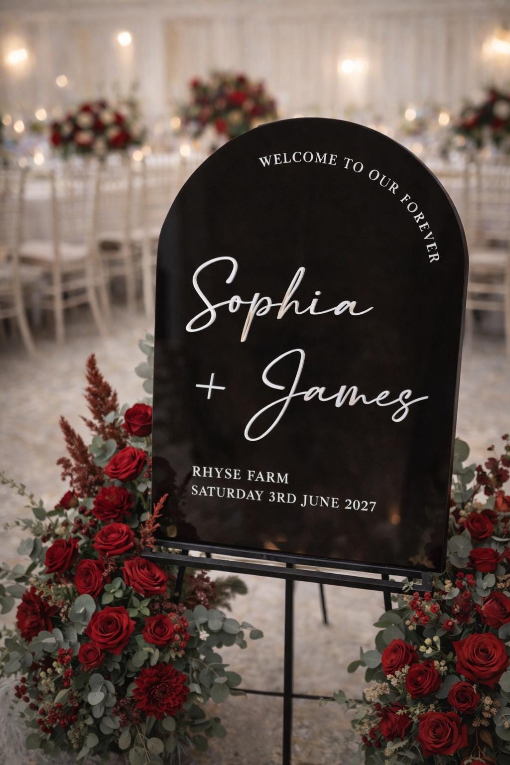 a black sign with white text welcoming guests to a wedding reception. The sign is surrounded by red flowers, and there are chairs and tables visible in the background, suggesting a wedding venue.