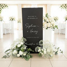 a black sign with white text welcoming guests to the wedding of Sophia and Harry, surrounded by white flowers and greenery.
