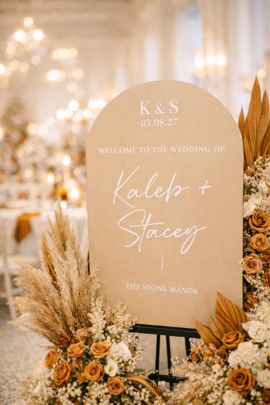 a wedding welcome sign with the names "Kaleb + Stacey" and the date "03.08.27" displayed prominently. The sign is surrounded by a floral arrangement, creating a warm and inviting atmosphere for the wedding ceremony.