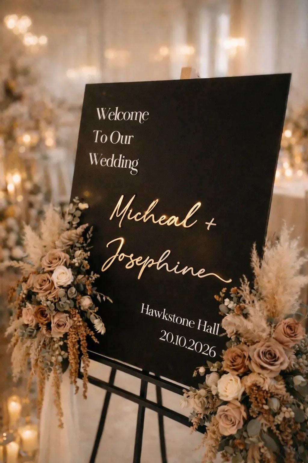 a black sign with gold lettering welcoming guests to a wedding. The sign is decorated with flowers and has the names "Michael + Josephine" written on it. The background of the image is blurred, but it appears to be a wedding reception venue with lights and other decorations.