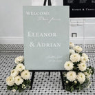 A wedding welcome sign with the names 'Ella' and 'Adrian' on it, surrounded by white flowers.