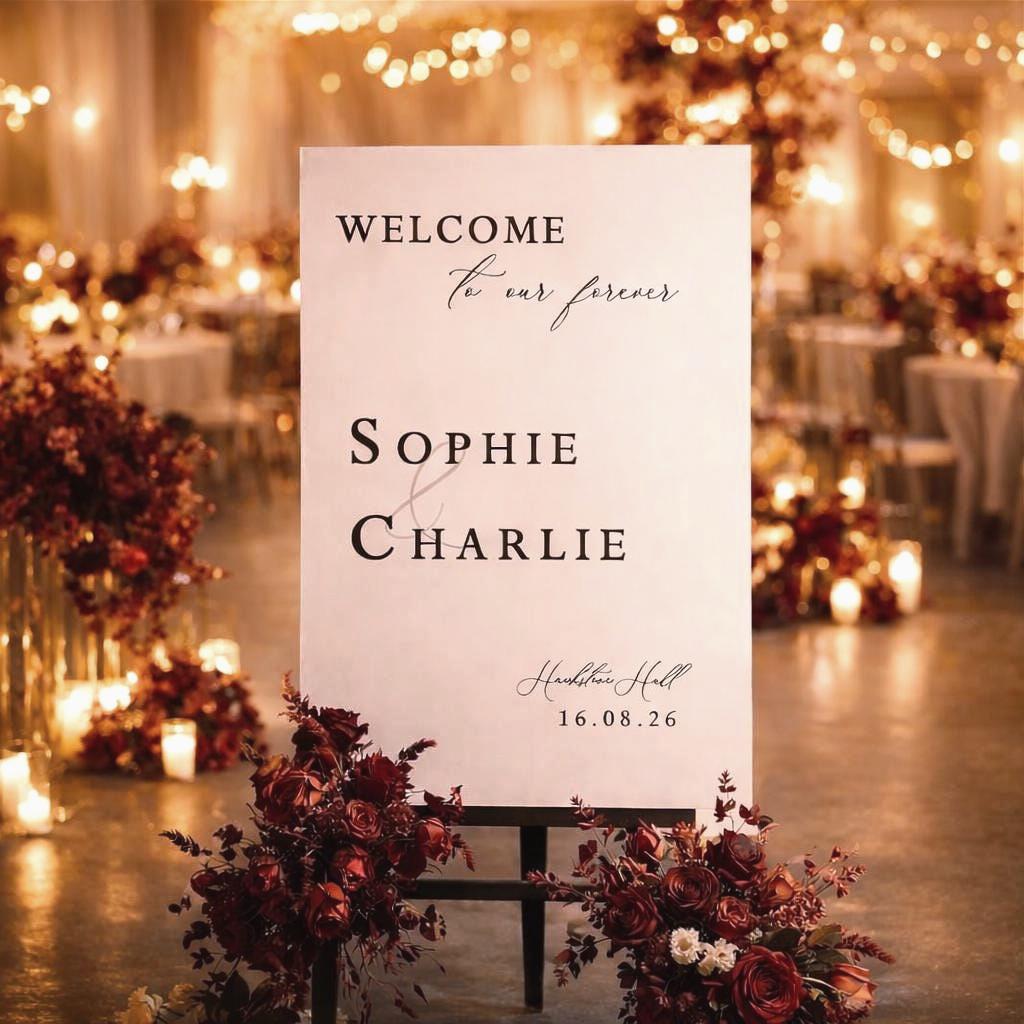 a wedding sign that reads "Welcome to our forever Sophie Charlie" with the date "16.08.26" below it. The sign is surrounded by red flowers and is placed in a room with a warm, festive atmosphere created by the lights and decorations.
