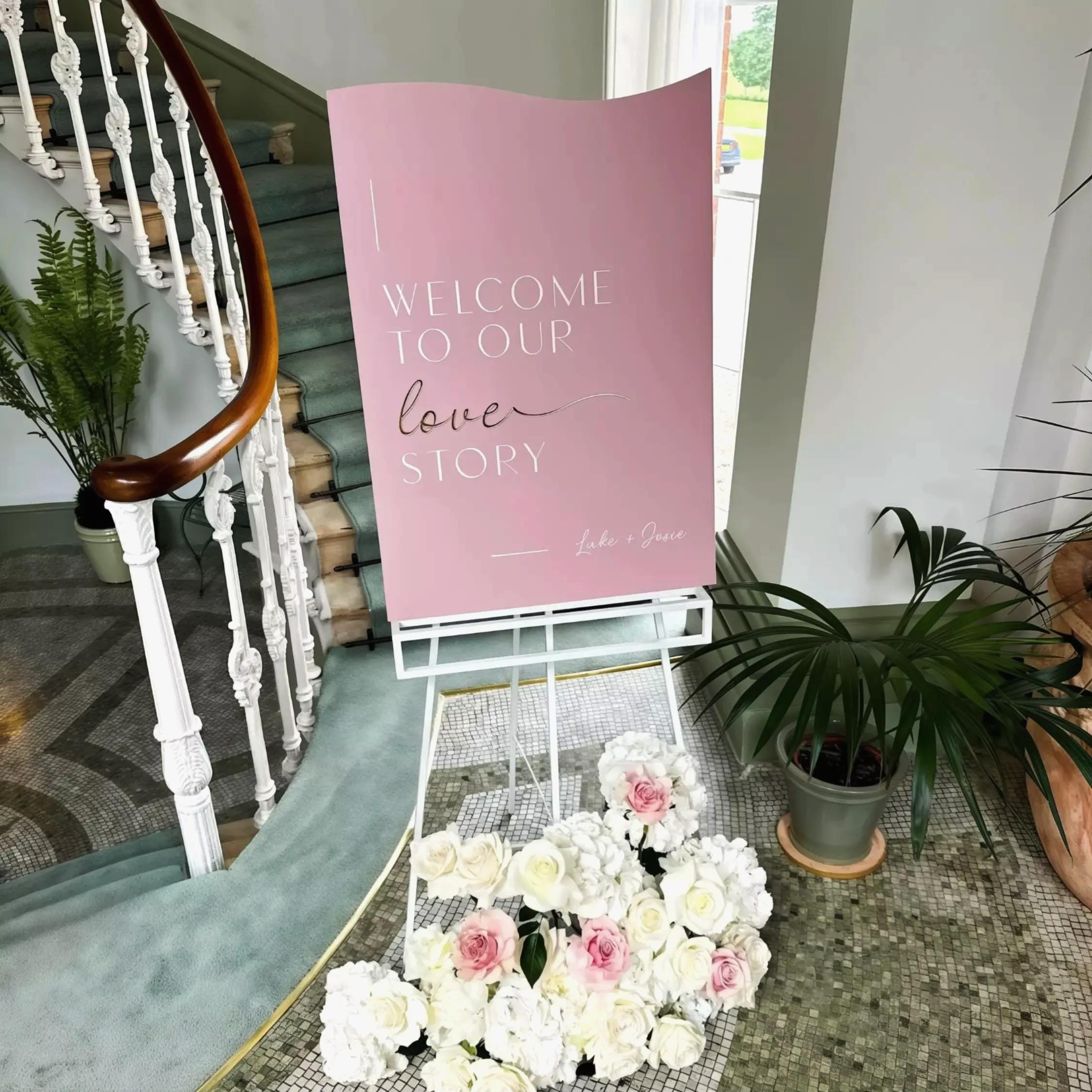A pink sign welcoming guests to a wedding, with a bouquet of white and pink flowers in front of it.